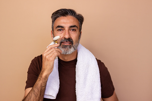 Confident middle-aged man with towel over shoulder using a facial roller