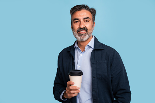 Smiling middle-aged man in casual wear holding a takeaway coffee cup, isolated on blue