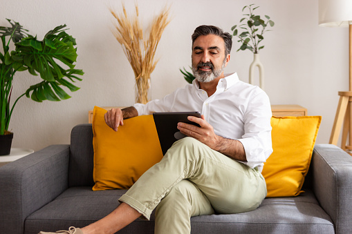 Confident mature male uses a digital tablet comfortably on his sofa, displaying technology use at home