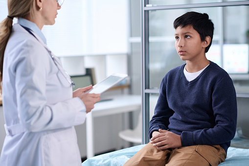 Portrait of young Middle Eastern boy talking to doctor and sitting on cot during consultation in clinic