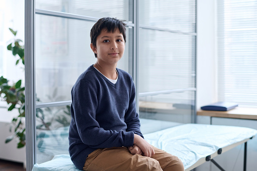 Portrait of young Middle Eastern boy sitting on cot in medical clinic and looking at camera with smile copy space