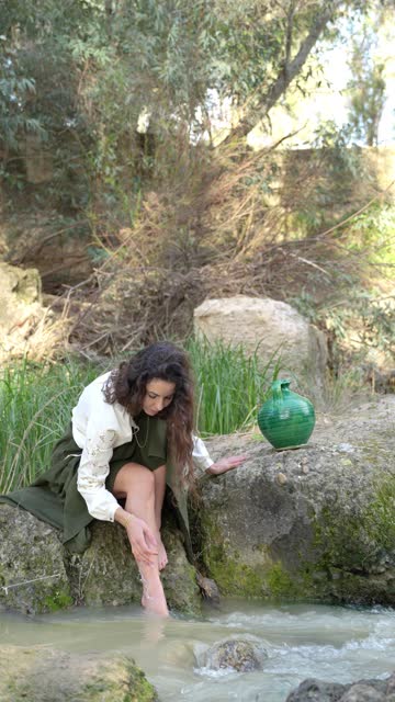 Woman plays with the flowing water of a river next to a green ceramic jug outdoors in nature.