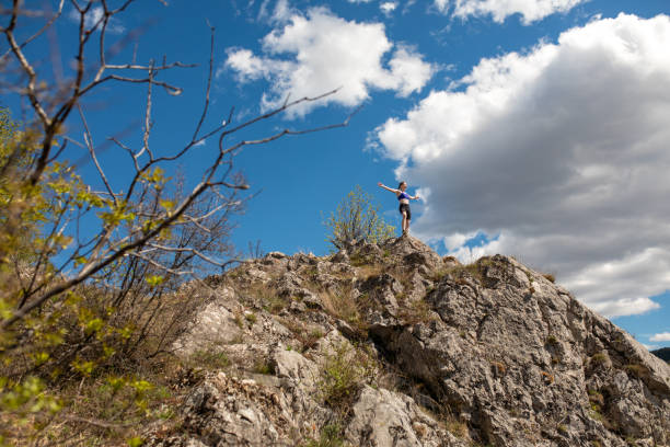 Man wearing helmet and harness looks up at rock face with climbing rope over shoulder. Outdoor rock climbing gear laid out on rocky terrain. Mountain adventure and climbing preparation.