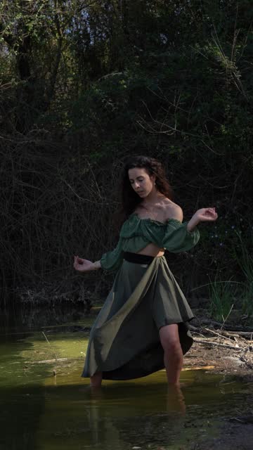 Young woman dances within the river's water, performing a rhythmic and tribal dance, fully immersed in the natural surroundings.