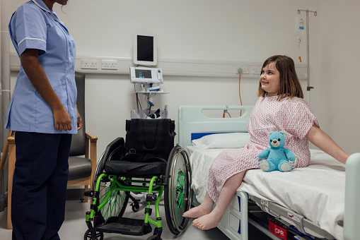 A shot of a young girl sitting on a hospital bed, wearing a gown during a routine visit to a hospital in Newcastle upon Tyne, North East England. She is paralysed from the waist down following a car accident when she was a baby. She is smiling up at the unrecognisable nurse who is standing opposite her and there is a teddy bear beside her on the bed. Her wheelchair is stationed next to the bed.
