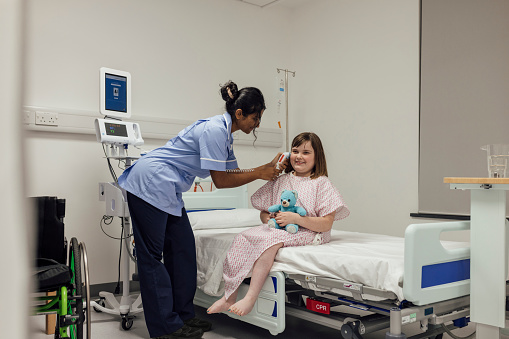 A shot of a young girl sitting on a hospital bed, wearing a gown during a routine visit to a hospital in Newcastle upon Tyne, North East England. She is paralysed from the waist down following a car accident when she was a baby. She is smiling and holding her teddy bear, whilst a nurse takes her temperature using an electronic thermometer in one of her ears. The young girl's wheelchair is stationed next to the bed, in partial view.