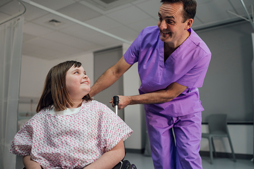 A shot of a young girl in a wheelchair wearing a hospital gown, smiling with a male doctor who is standing beside her in a hospital in Newcastle upon Tyne, North East England. The young girl is paralysed from the waist down following a car accident when she was a baby and is visiting hospital for a routine check-up. The doctor is holding onto the handles of her wheelchair.