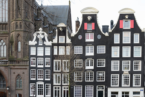 Brown facades of buildings in old Amsterdam. White windows. Gable roofs are decorated with ornaments. Part of the cathedral is visible on the left.