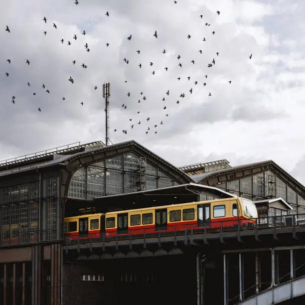 Industrial landscape, a vibrant yellow and red train stands on the railway station. Birds flock and the cloudy sky over the metal urban architecture
