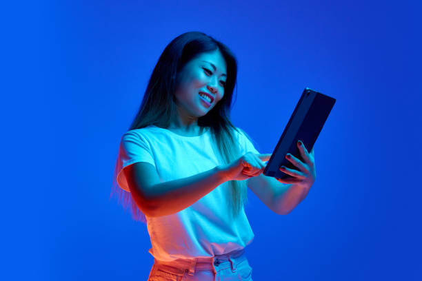 istock Young, attractive Asian woman using digital tablet against blue gradient background in neon light. Working in a distance. 2147712035