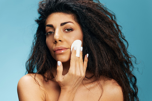 Tanned pensive curly Latin female holding cotton pad for face, cleansing face with toner after shower over blue studio background. Happy woman enjoying fresh moisturized face skin. Copy space