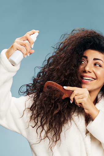 Hairstyling. Enjoyed tanned curly Latin lady in bathrobe Spraying On Hair For Repair split ends posing isolated on pastel blue background, using hairbrush, looking up. Hair Care Ad Concept offer