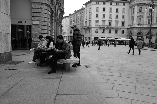 Milan, Italy - April 09, 2024: some people on foot  are crossing Piazza del Duomo in Milan. the people are sitting on the bench