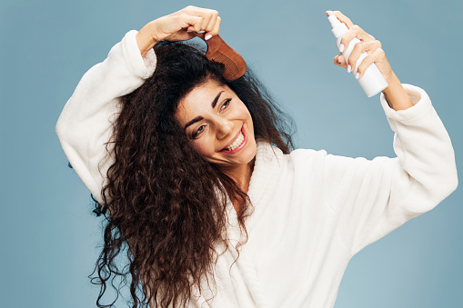 Hairstyling. Smiling tanned curly Latin lady in bathrobe Spraying On Hair For Repair split ends posing isolated on pastel blue background, using hairbrush, looking aside. Hair Care Ad Concept offer