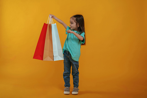 Shopping bags in hand. Cute little girl is against yellow background.