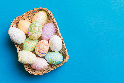Colorful Easter eggs in wicker basket against colored background, closeup. top view with copy space.