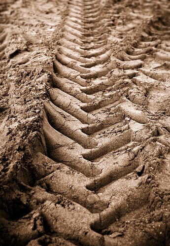 Vertical photograph of brown muddy field or mud ground land patch or dirt or sand gravel pitch with tyre markings design, tire treads or grooves or patterns with shallow large big marks making small pits.