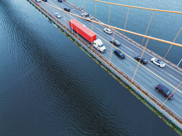 Aerial view of a semi truck with red trailer crosses a suspension bridge.
