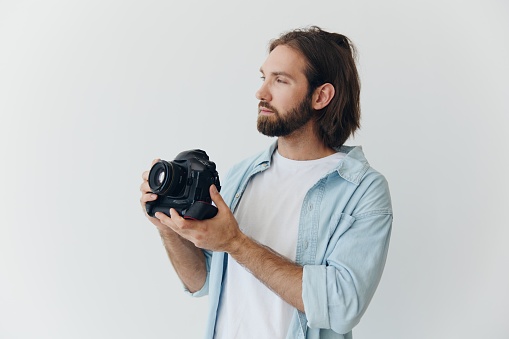 A male hipster photographer in a studio against a white background looks through the camera viewfinder and shoots shots with natural light from the window. Lifestyle work as a freelance photographer. High quality photo
