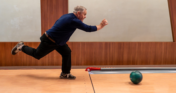 Senior Man Throwing Ball in Bowling Game. Energetic elderly man in action, bowling at recreational facility with enthusiasm.