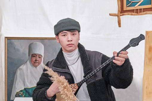 Almaty, Kazakhstan - March 21, 2024: Teenager playing dombra in tent with souvenirs and looking at camera during spring festival Nowruz. Kazakh folk two-stringed musical instrument. National music.