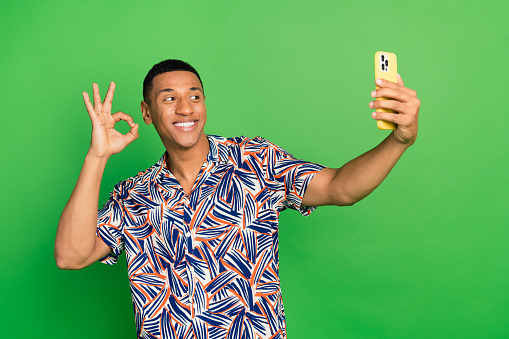 Photo of cheerful funny guy stylish summer clothes shooting filming looking camera webcam okey sign isolated on green color background.