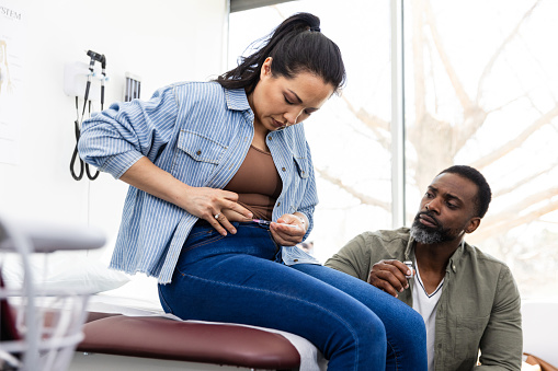 The mid adult husband watches as his mid adult wife injects the medication into her abdomen with a syringe.