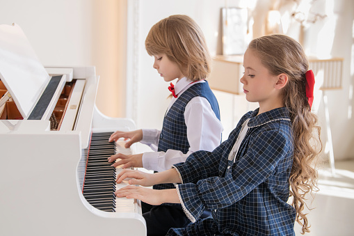 Two children are playing the piano. One is wearing a blue shirt and the other is wearing a blue vest