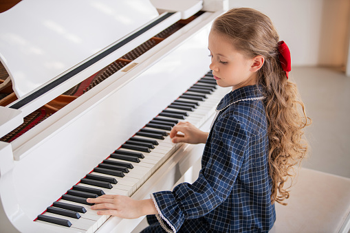 A young girl is playing the piano. She is wearing a blue shirt and a red bow. The piano is white and has black keys