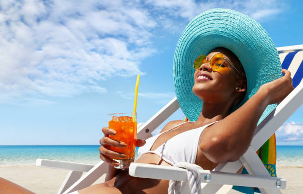 la mujer feliz está tomando el sol en una tumbona de playa, con sombrero para el sol y gafas de sol, bebiendo un jugo de naranja en un día soleado junto al mar, concepto de unas vacaciones de verano en la playa, reserva de viajes y resort - caribbean sea fotografías e imágenes de stock