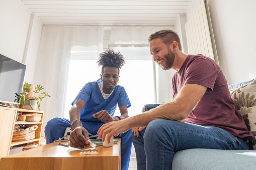 A healthcare professional in blue scrubs attentively providing medication to a patient in a well-lit, comfortable home setting. The patient, seated on a blue couch and wearing a casual maroon shirt with a flower decorated pillow against him. He is interacting with the healthcare worker. They are surrounded by a tidy living room, with a wooden coffee table where medical supplies, a tablet, and a bottle of medication are neatly arranged. The soft natural light from the window creates a calming atmosphere, complemented by the light sheer curtains and the homey decor, including a flat-screen TV and various plants that add a touch of greenery to the room.