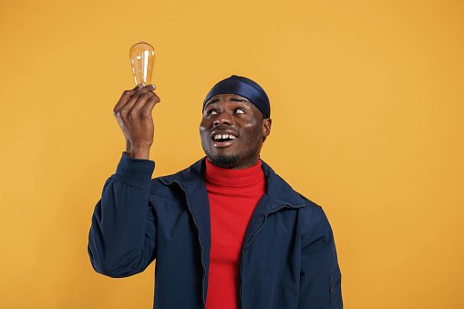 Light bulb in hand, having an idea. Handsome black man is in the studio against yellow background.
