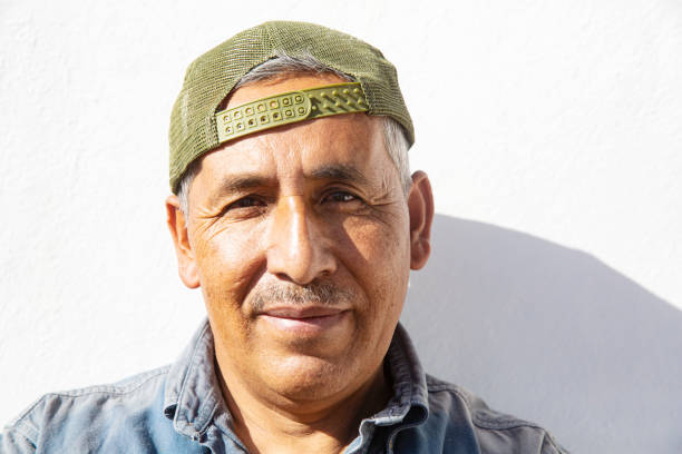 A middle aged man with a strong face, wearing a backwards baseball cap, against a white background lit by strong sunlight