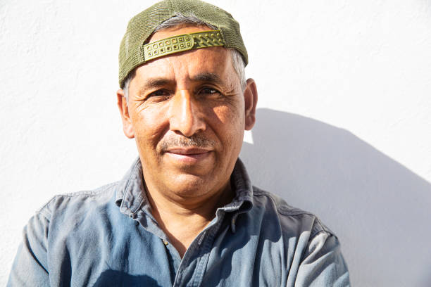 A middle aged man with a strong face, wearing a backwards baseball cap, against a white background lit by strong sunlight