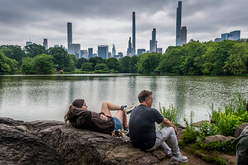 A couple enjoys the fine weather in Central Park sit or climb on Hern Point, an outcropping of Manhattan Schist on the west side of the Boat Lake. In the background to the south is the skyline of Central Park South and “Billionaire’s Row” on 57th Street.
