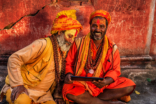 Sadhu - indian holymen using the digital tablet. In Hinduism, sadhu, or shadhu is a common term for a mystic, an ascetic, practitioner of yoga (yogi) and/or wandering monks. The sadhu is solely dedicated to achieving the fourth and final Hindu goal of life, moksha (liberation), through meditation and contemplation of Brahman. Sadhus often wear ochre-colored clothing, symbolizing renunciation.