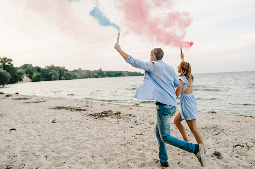 Man and woman standing and kissing on sand and holding colorful blue and red, pink color smoke bombs on beach. A happy married couple with a smoke bomb. Baby shower. Twins: boy and girl. Side view
