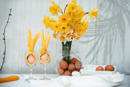 Egg wrapped in bunny ears Yellow napkin in wine glasses and bouquet of Daffodil flowers in glass vase with boiled eggs for Easter Holiday