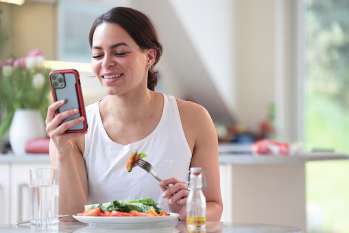 Multiracial woman uses phone while eating salad at home