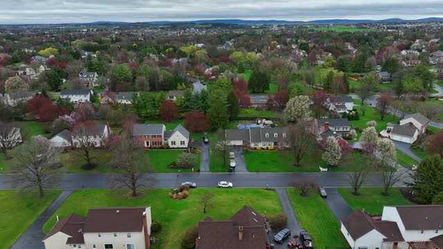 Lush green grass and colorful trees during spring season. Rainy day in american residential suburb. Noble one family homes and villas with private swimming pools. Aerial lateral wide shot. PA, USA.