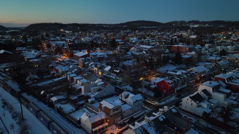 Train on track in small american town during winter snow. Sunset time at horizon over lighting buildings and homes. Snow covered roofs in illuminated city of USA. Aerial forward flight. Wide shot.