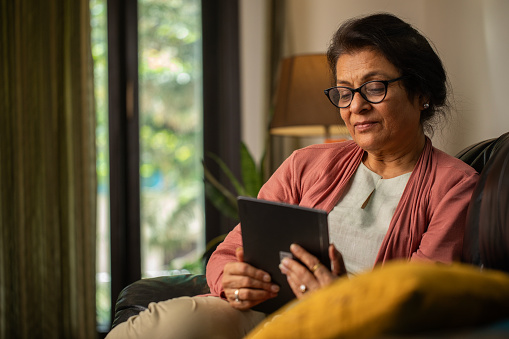 Focused senior woman in eyeglasses watching movie over digital tablet while relaxing on sofa in living room