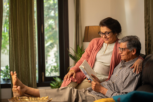 Smiling senior couple in eyeglasses reading book while sitting together on sofa in living room at home