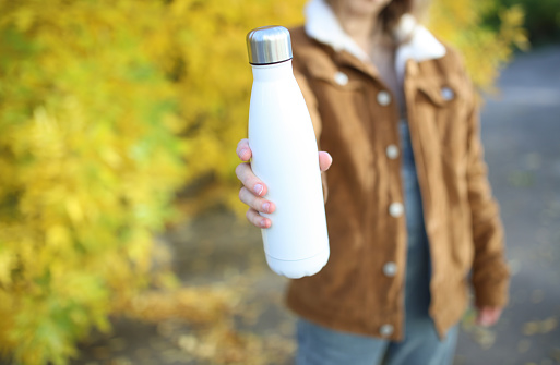 Woman holding a white thermos bottle against the background of autumn trees