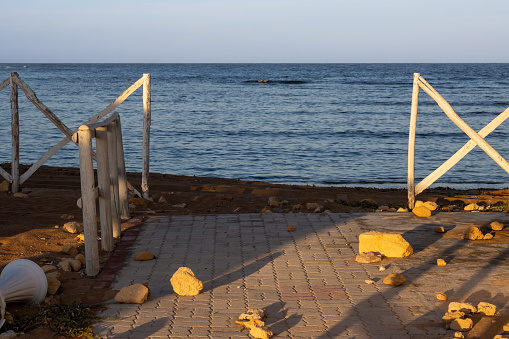 Calm water of the Mediterranean sea. White wooden fence, colored by morning sunshine to a golden color, together with the stones. Spiaggia Sibilliana, Marsala, West Sicily, Italy.