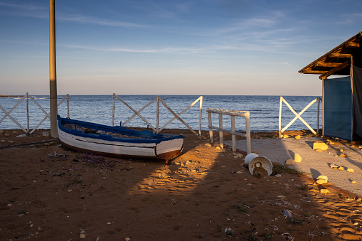 Calm water of the Mediterranean sea. White wooden fence, small beach house and a wooden boat in the sand. Spiaggia Sibilliana, Marsala, West Sicily, Italy.