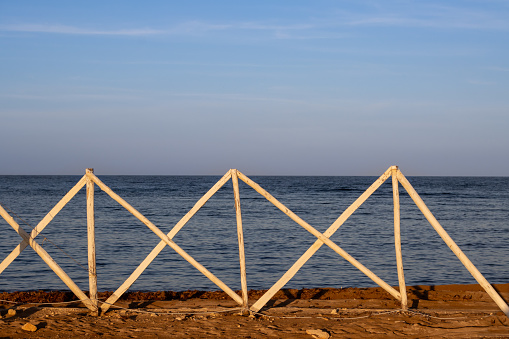 Calm water of the Mediterranean sea. White wooden fence, colored by morning sunshine to a golden color, together with the stones. Spiaggia Sibilliana, Marsala, West Sicily, Italy.
