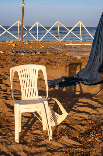 Outdoors still life. Scene in a morning sunlight, coloring to golden a white beach chair and a wooden fence and Mediterranean sea. Dark blue drapery. Spiaggia Sibilliana, Marsala, West Sicily, Italy.