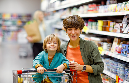 A smiling woman and her young son select products while pushing a shopping cart in the supermarket, showcasing their engaging family activity.