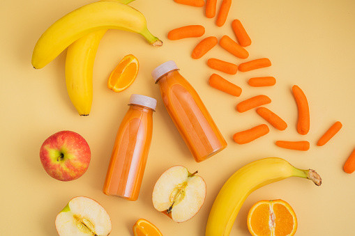 Two bottles of freshly made smoothie or orange juice alongside bananas, apples,  carrots, and oranges on a yellow background. Nutrition and wellness concept.